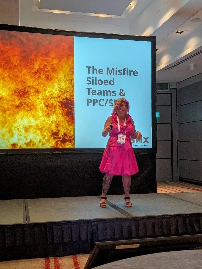 Amanda Friedt presenting on the SMX main stage in a hot-pink dress, a wall of flame on the screen behind her beside the talk title 'The Misfire: Siloed Teams & PPC/SEO'.